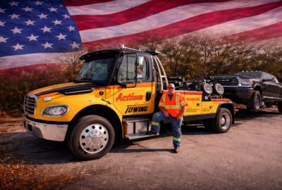 Tow truck with a worker in an orange safety vest standing beside a yellow Action Towing truck, carrying a car on a flatbed with an American flag backdrop.