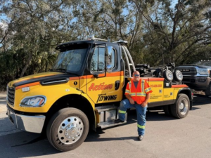 Yellow Action Towing tow truck parked on a road with a driver wearing an orange safety vest leaning against the cab.