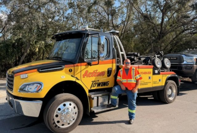 Yellow Action Towing tow truck parked on a road with a driver wearing an orange safety vest leaning against the cab.